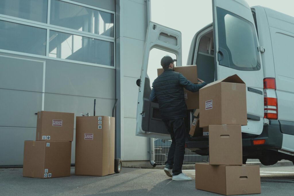 A delivery man unloading cardboard boxes from a van at a warehouse during the day.